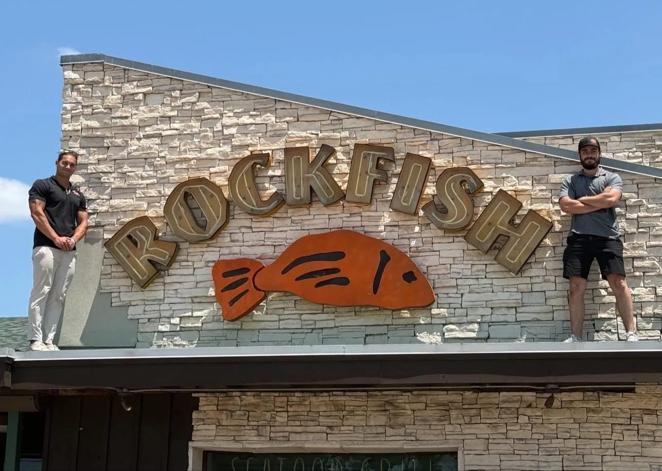 Two TDC crew members standing on the roof of a stone-fronted restaurant, either side of a large Rockfish sign