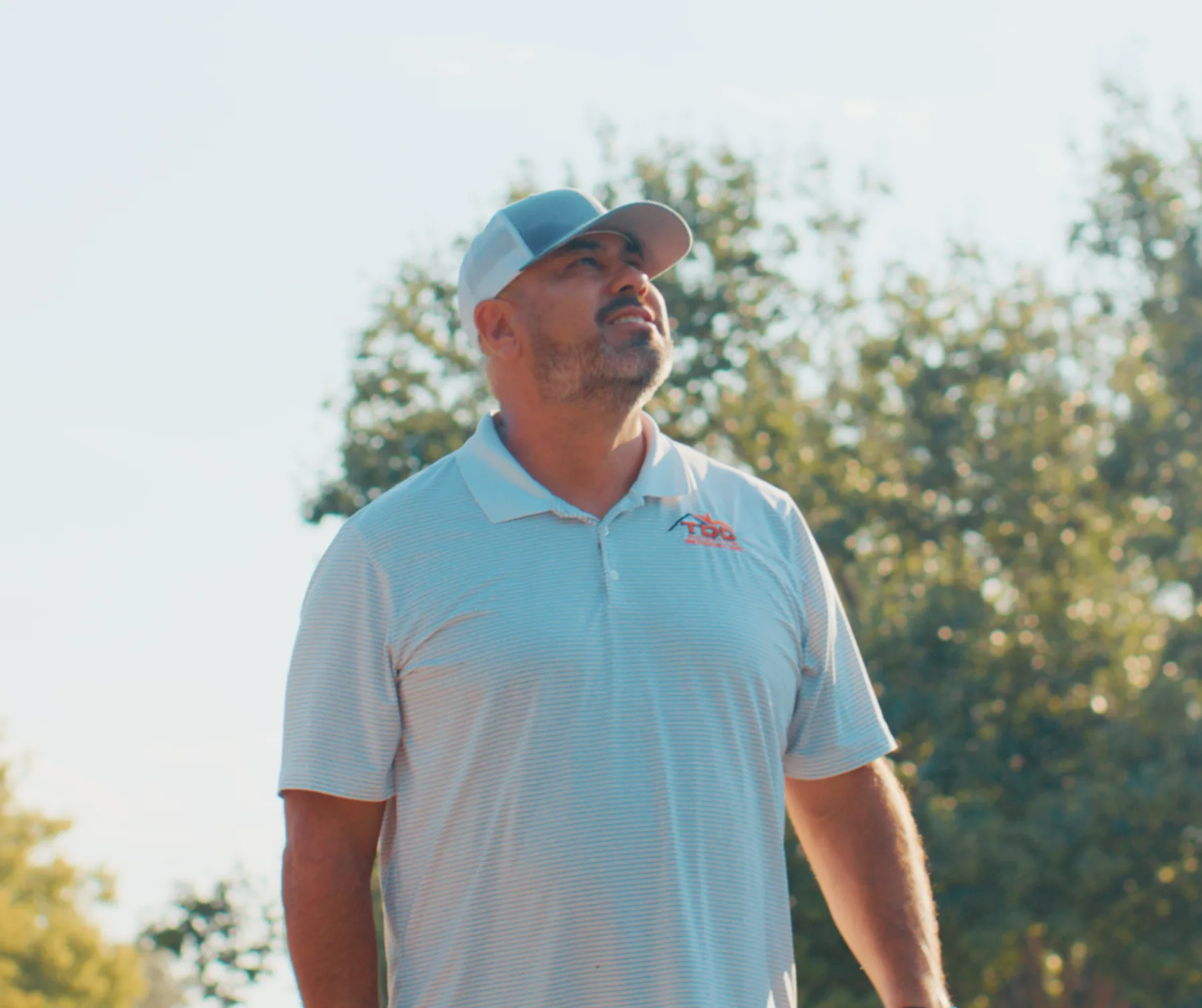 David Carrasco, owner of TDC Roofing and Remodeling, in a branded polo shirt and cap, looking up at a roof