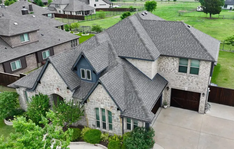 Drone view of a new gray shingle roof on a two-story stone home with a navy gabled entry, seen from above the driveway