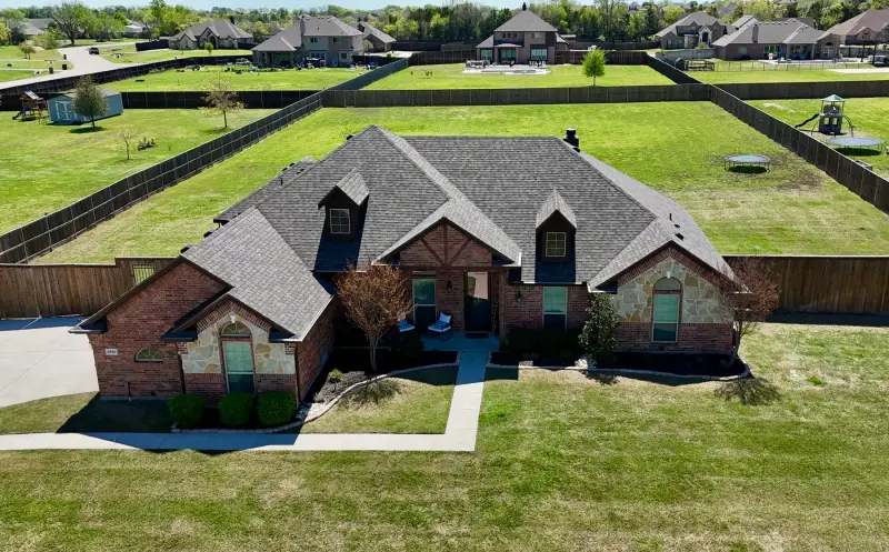 Drone view from the front of a brick and stone home with a new gray shingle roof and two dormers, on a large fenced lot