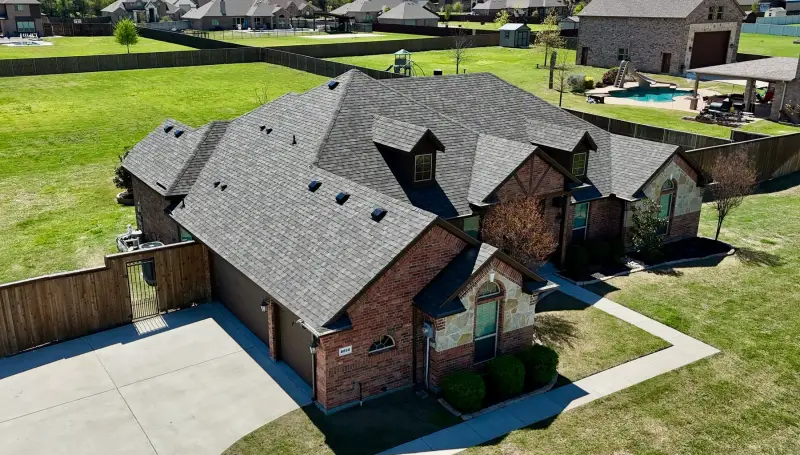 Drone view of a new gray shingle roof on a single-story brick home with a three-car garage, a fenced yard and a pool behind it