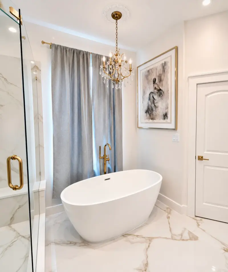 Remodeled bathroom with a white freestanding tub, a gold floor-mounted filler, a chandelier, marble-look tile and a gray curtain