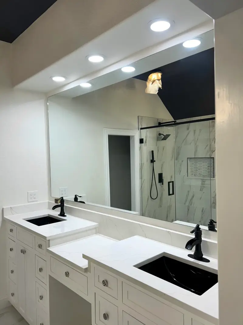 Remodeled bathroom vanity with white cabinets, a white counter, a black faucet and a wide mirror reflecting the glass shower with black fixtures