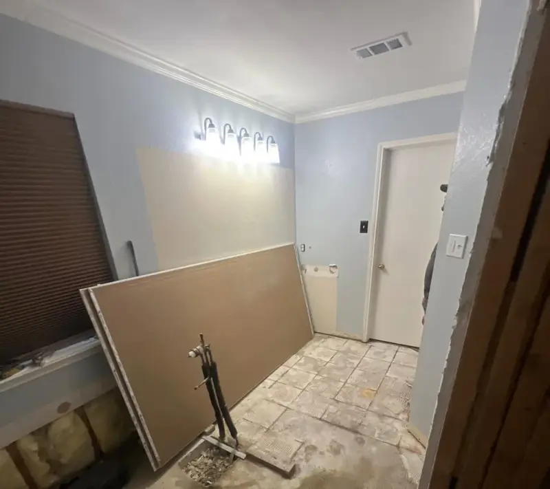The same bathroom mid-demolition, with the vanity removed, the floor tile stripped, a sheet of drywall leaning on the wall and the old light bar still up