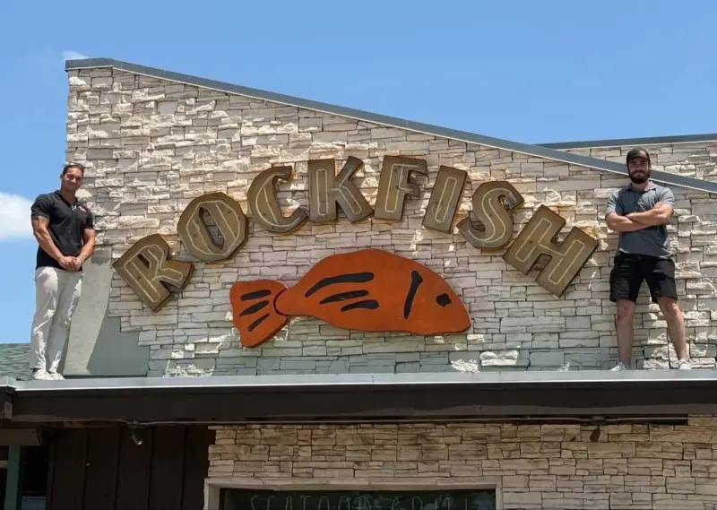 Two TDC crew members standing on the roof of a stone-fronted restaurant, either side of a large Rockfish sign