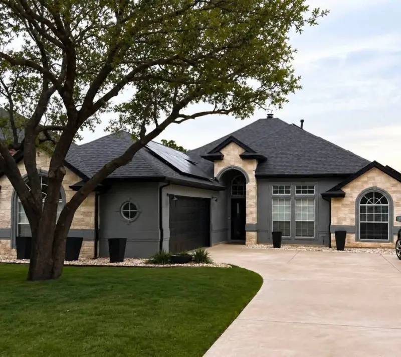 Single-story home after an exterior remodel, with the brick painted gray, black trim, a black garage door and a new dark shingle roof under a large oak