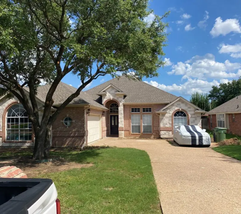 The same home before, with tan brick, cream trim, a white garage door and a faded gray roof