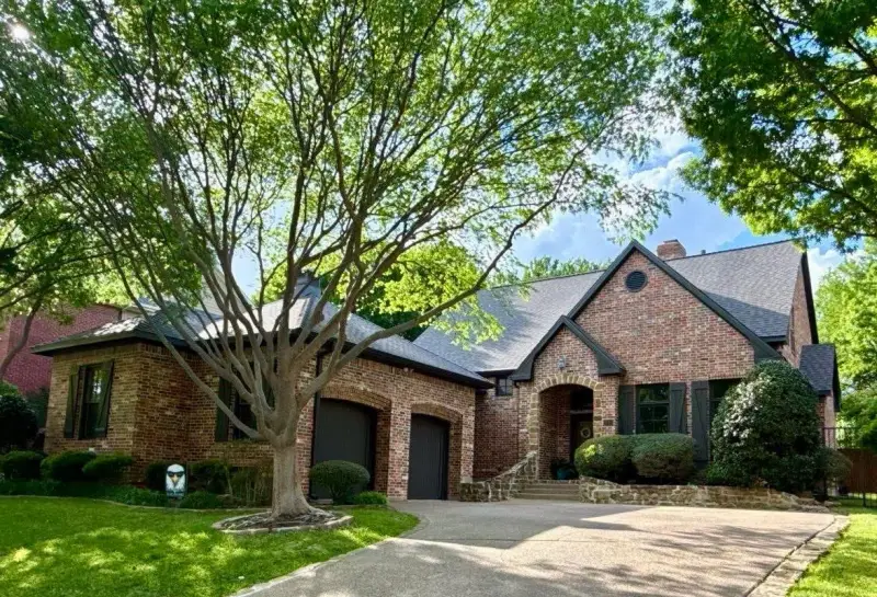 Street view of a red brick home with a dark shingle roof and two garage doors, framed by a large tree in the front yard