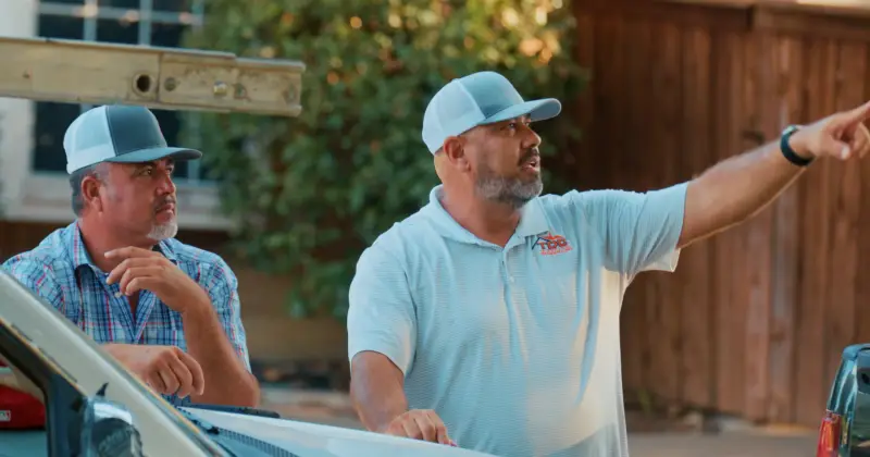 A TDC representative in a branded polo pointing up at a roof while explaining the work to a homeowner beside a truck