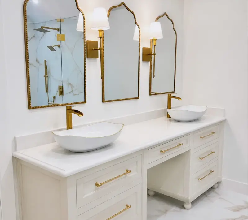Finished bathroom vanity with a white marble counter, two vessel sinks, gold faucets and sconces, three arched gold mirrors and cream cabinets on marble floors