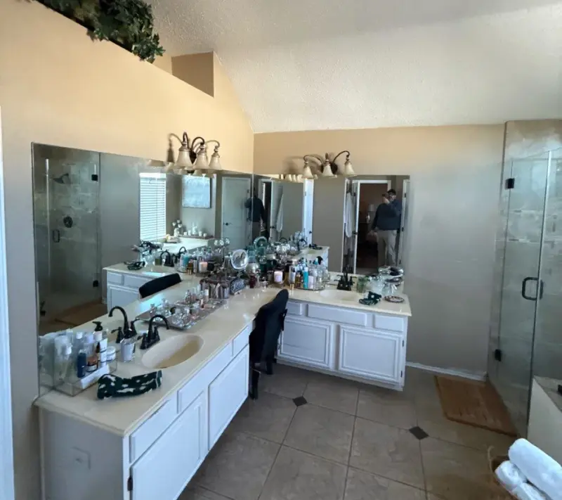 The same bathroom before the remodel, with a cream tile counter, oval drop-in sinks, oak-trimmed mirrors and beige tile floors