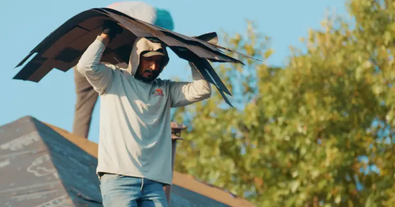 A TDC roofer in a branded shirt carrying a bundle of shingles across his shoulders on a roof against a blue sky