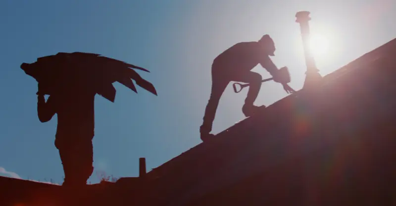 Two roofers silhouetted against a low sun on a roof, one carrying shingles and one working along the ridge