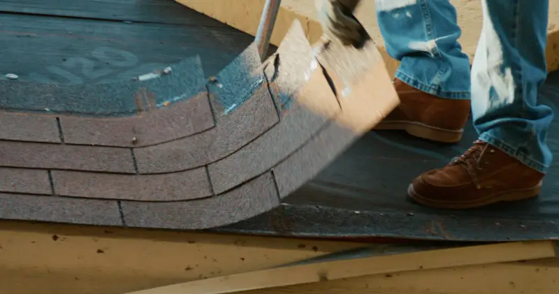 Close view of a roofing shovel prying up a row of old shingles and nails, with a roofer's boots braced on the deck
