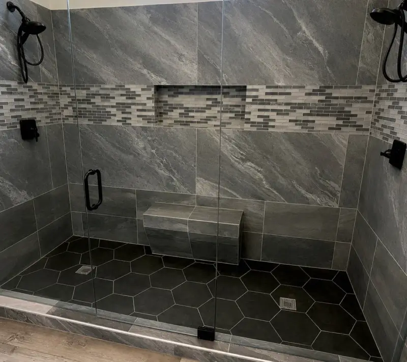 Finished walk-in shower with large gray stone-look tile, a mosaic accent band and niche, a tiled bench, hexagon floor tile, two black shower heads and a frameless glass door