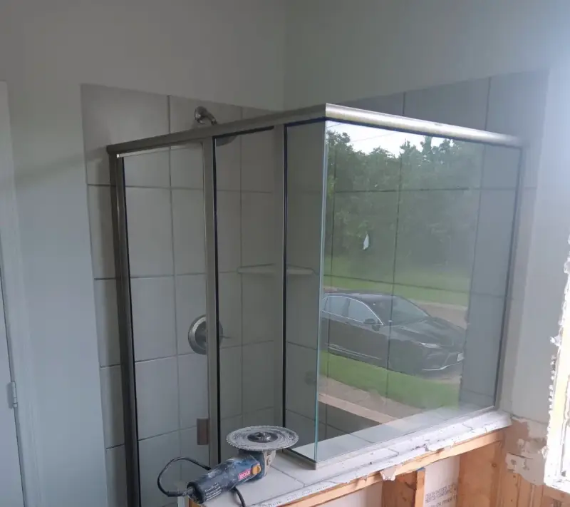 The old shower during demolition, with white square tile, a framed glass enclosure and a grinder on the sill where the wall was opened up